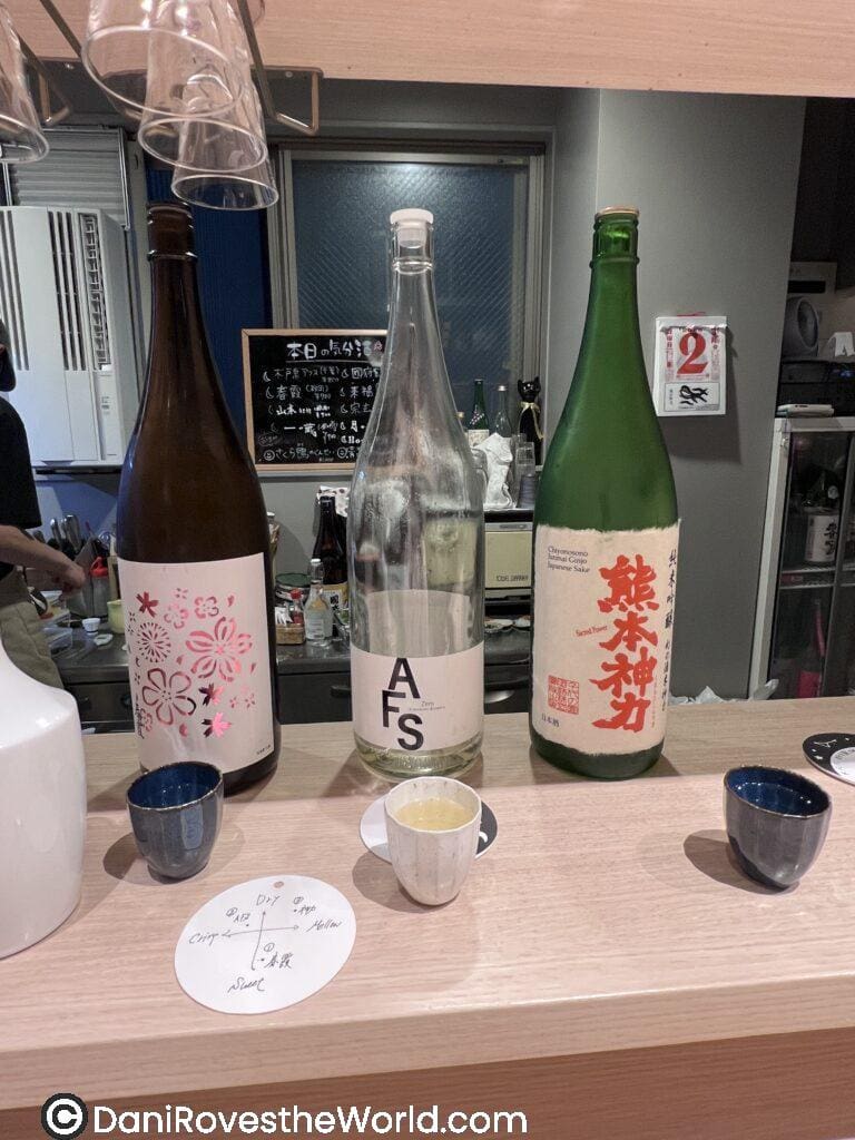 3 bottles of sake on a counter. one bottle has pink flowers, the middle bottle has the initals AFS and the right bottle has japanese letters in red