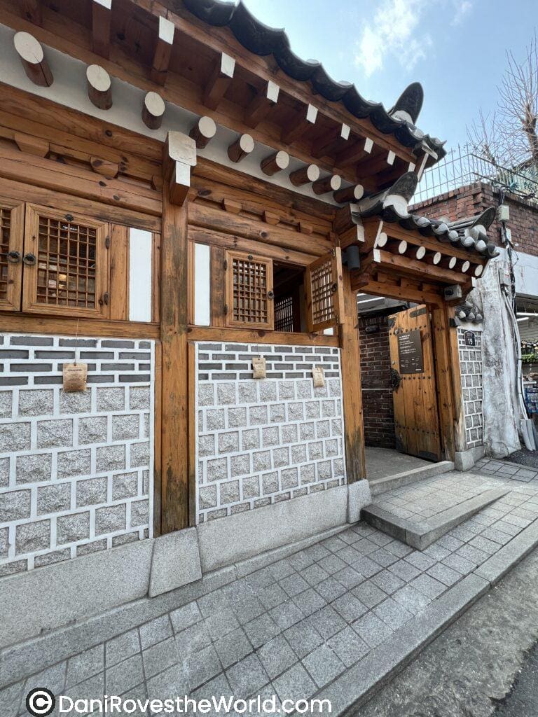 Bukchon Hanok Village in Seoul.: A traditional Korean hanok entrance with wooden beams, tiled roof, and small lattice windows. The stone wall is patterned with square blocks and accented by wooden pillars.