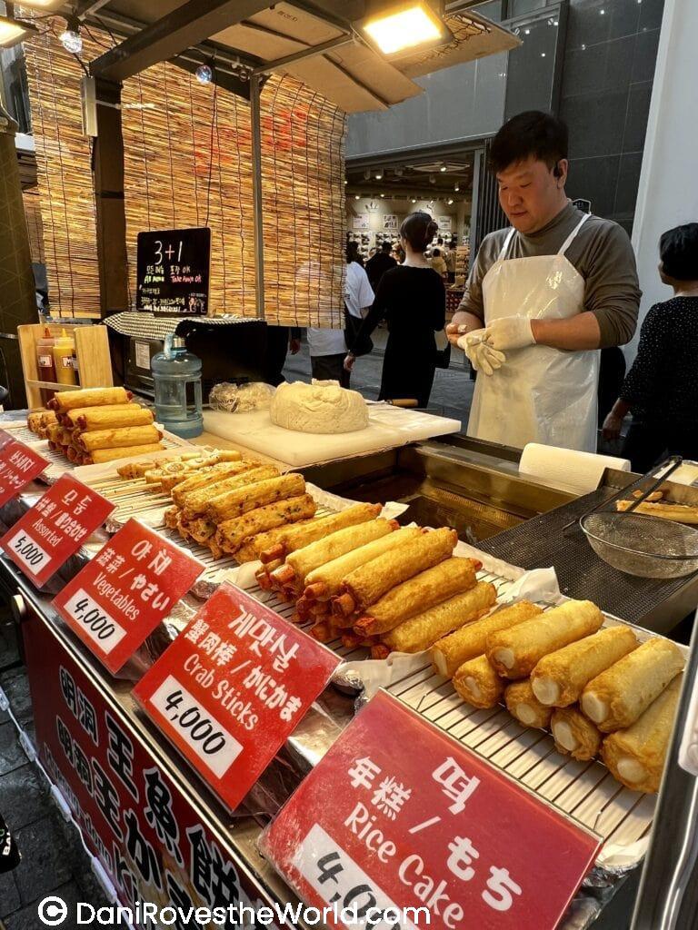 A food stall in Myeongdong selling various fried snacks like crab sticks and rice cakes, with bright red signs displaying prices. Trying street food is one of the top things to do in Myeongdong, with an array of delicious local treats to enjoy.