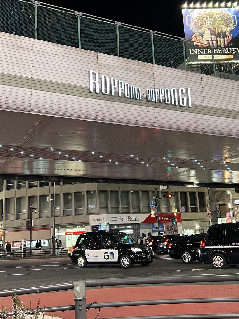 Street scene outside Roppongi at night with taxis and illuminated signage.