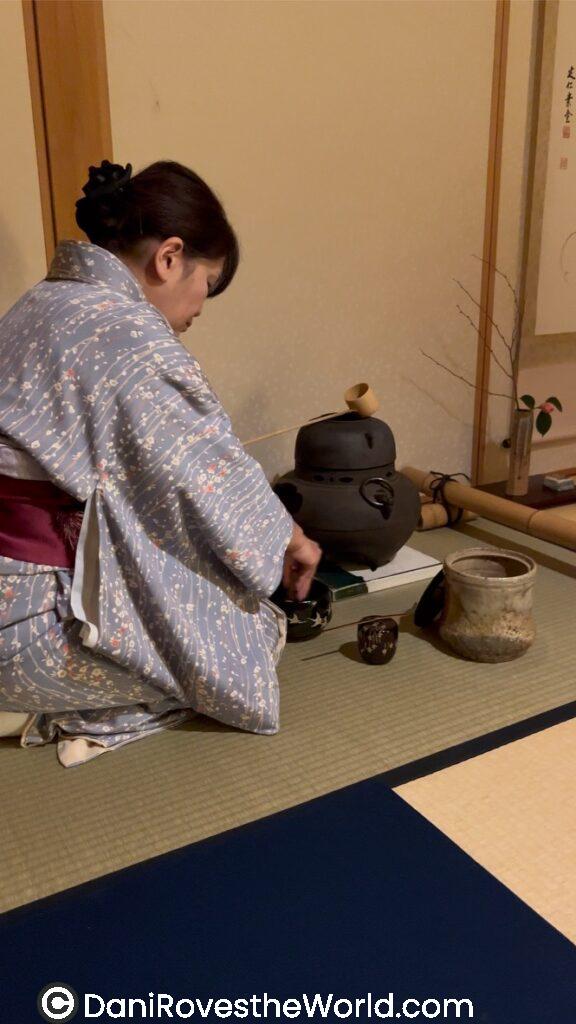 Japanese Tea Ceremony- a women wearing a kimono is kneeling in front of a black pot with a matcha bowl and other accessories 