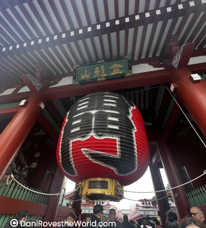 This image shows the iconic red paper lantern hanging at the entrance of Senso-ji Temple in Tokyo, Japan. The large lantern displays bold black kanji characters and is framed by vibrant red temple pillars, with visitors gathered below. Above the lantern is a green and gold plaque with additional kanji characters.