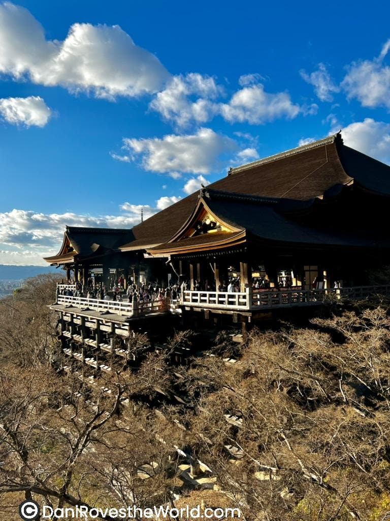 The historic wooden Kiyomizu-dera temple in Kyoto perched on a hillside, framed by bare tree branches under a vivid blue sky dotted with fluffy clouds.