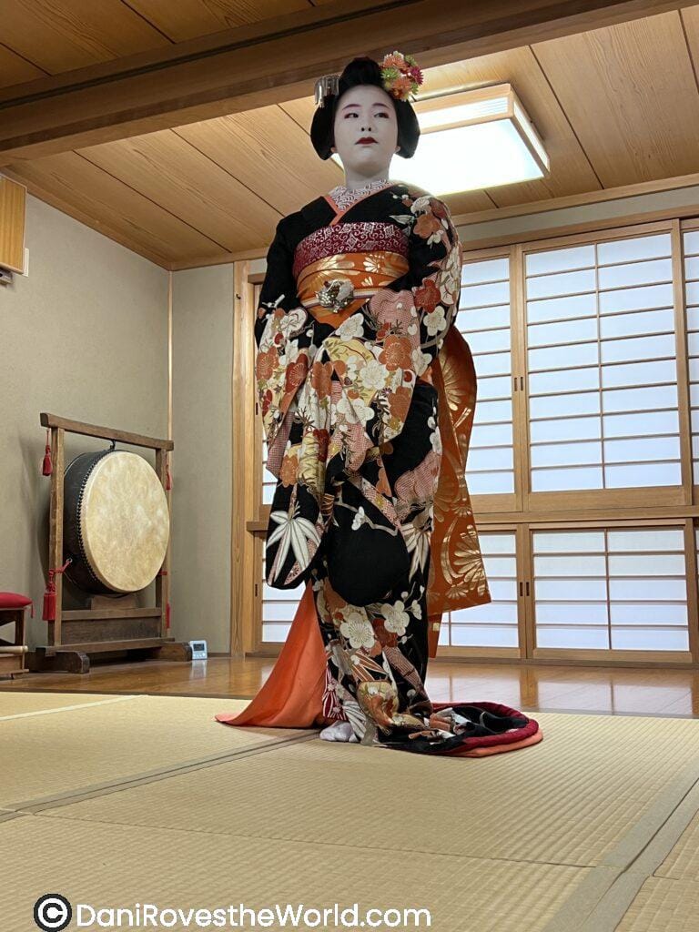 A Geiko showing off her traditional attire of kimono, custom flower headpiece and white makeup. This is a winter piece so colors are deep in reds, oranges and dark pinks.