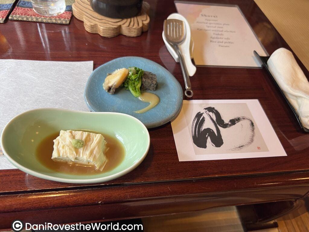 Kaiseki meal presentation: A traditional kaiseki meal on a lacquered wooden table, featuring yuba (tofu skin) in a light soy broth with wasabi, and a small plate of seasonal vegetables and tofu with miso sauce, alongside a printed menu and minimalist brush art.