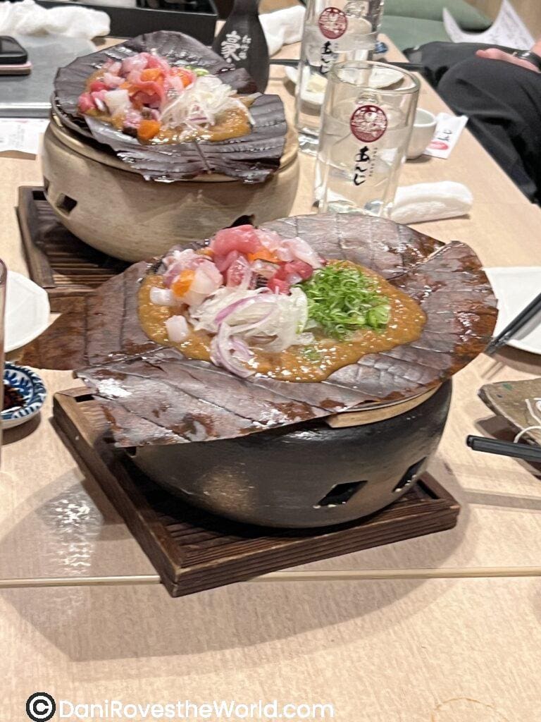 	3.	Hoba miso dinner dish: Hoba miso served on magnolia leaves over a clay grill, topped with chopped raw seafood, red onions, and scallions, part of a Kyoto izakaya meal experience, with sake glasses and tableware in the background.