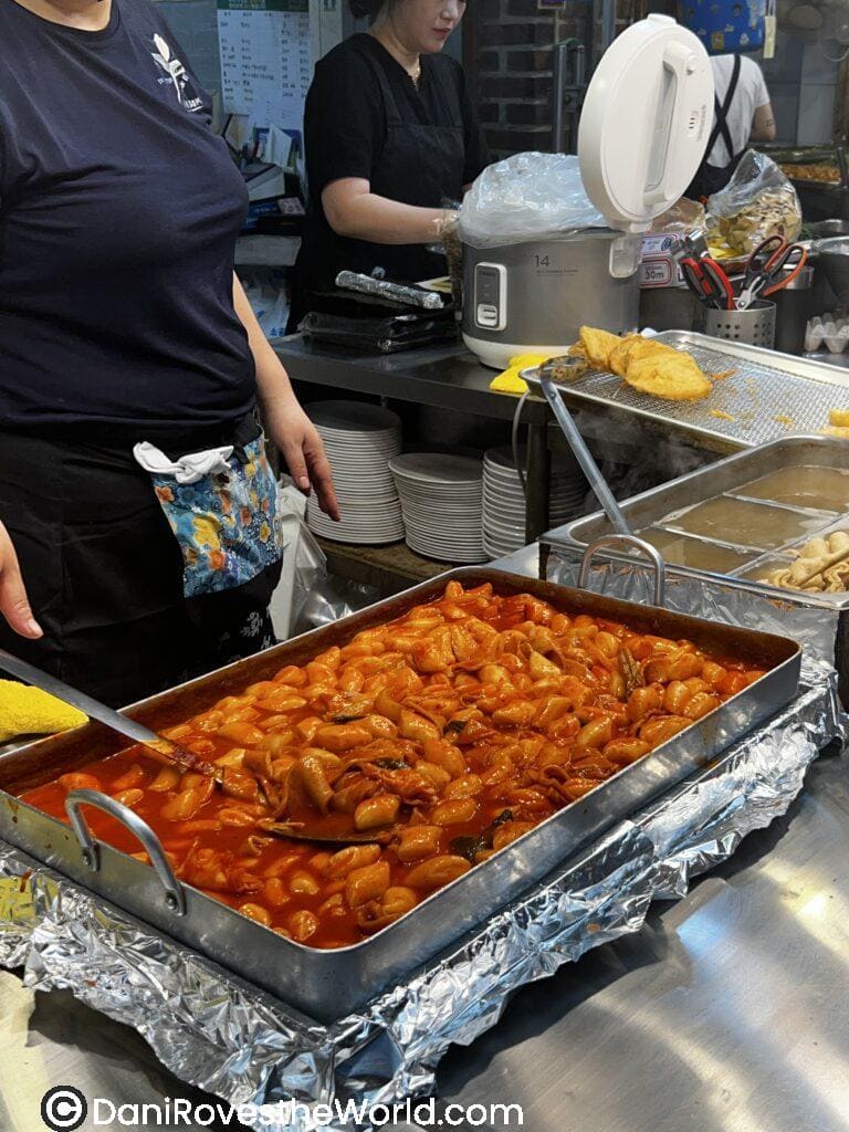 A tray of tteokbokki, a popular Korean street food made of rice cakes in a spicy red sauce, being served in a bustling Seoul food market.