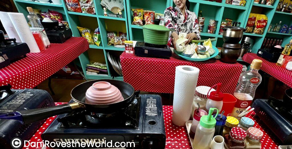Korean cooking class setup in Seoul with portable burners, ingredients, and a local instructor preparing vegetables in a colorful kitchen classroom.