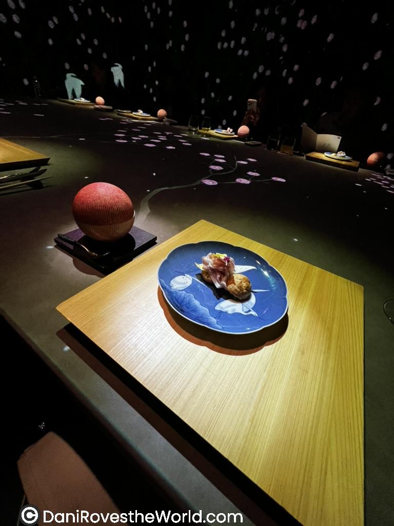 A beautifully plated dish at an upscale Tokyo restaurant, Moonflower Sagaya, featuring a small bite of intricately arranged sushi on a deep blue plate with white floral designs. The dimly lit setting is enhanced by soft, artistic lighting with delicate floral projections on the table, creating a serene and immersive dining experience.