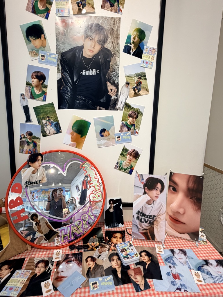 K-pop Birhday cafe fan display featuring photos, photocards, and merchandise arranged around a heart-shaped mirror with BTS lettering inside a themed café or shop interior in South Korea.