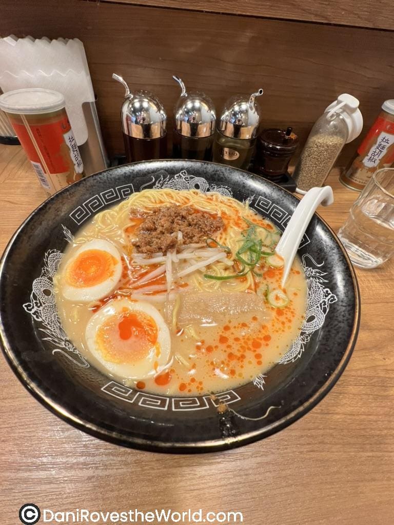 A steaming bowl of ramen served at a Tokyo ramen shop is one of the best tokyo eats for a budget meal! Featuring rich broth topped with soft-boiled eggs, ground pork, green onions, bean sprouts, and a drizzle of chili oil. The black bowl is adorned with intricate white patterns, while various condiments like sesame seeds and chili powder are available in the background for customization.