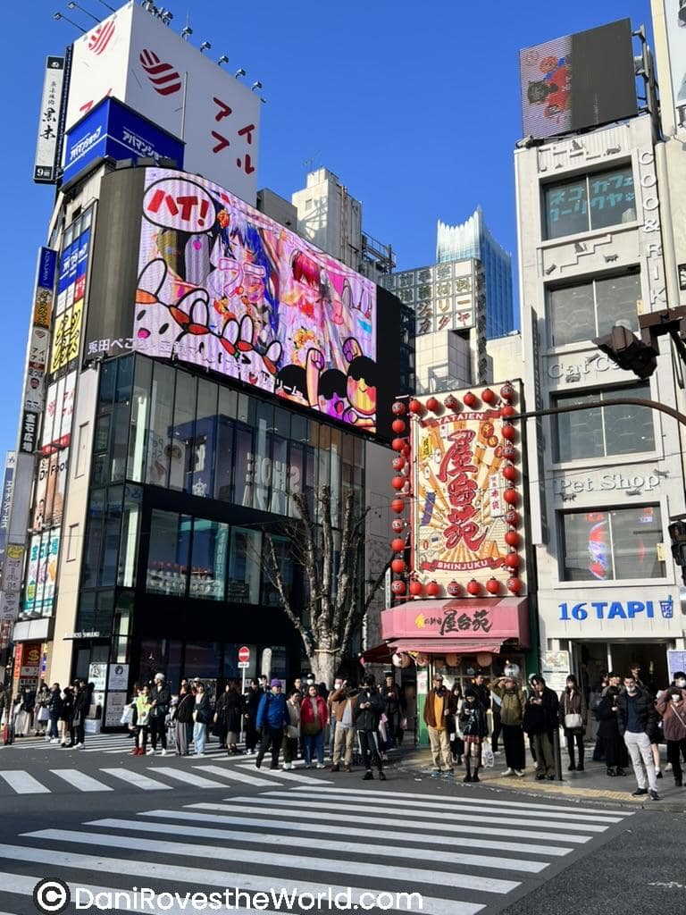 . Busy Shinjuku crossing filled with people under bright billboards and anime advertisements, highlighting a central spot for where to stay in Tokyo.