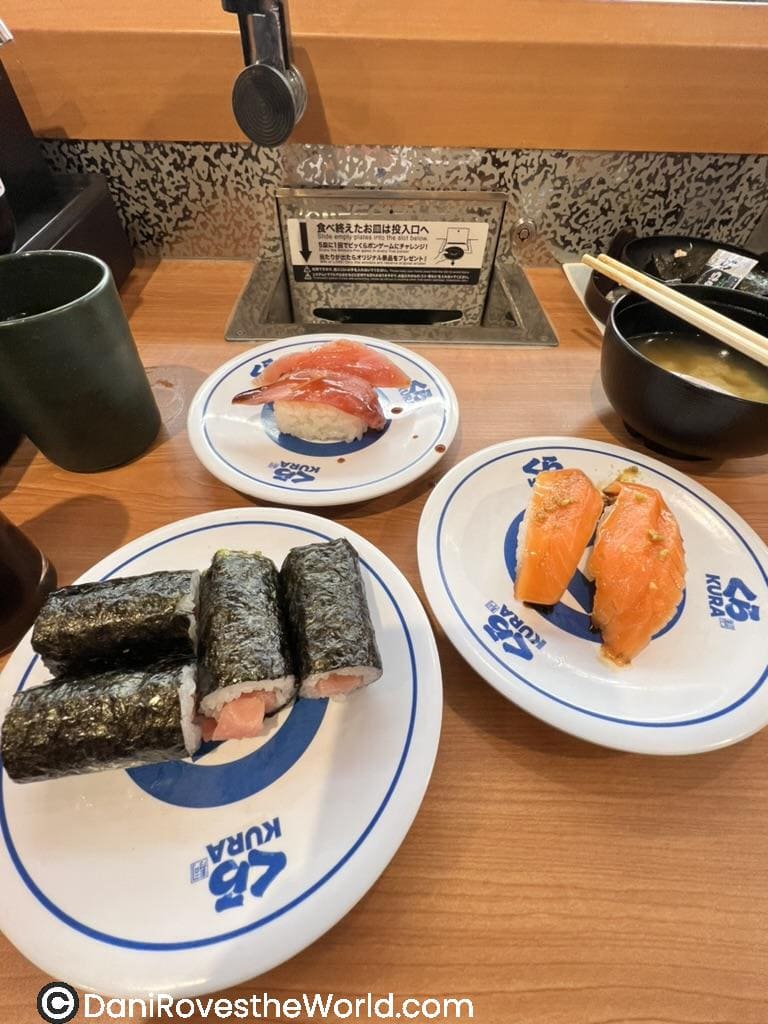 A variety of sushi dishes served at Kura Sushi, a popular conveyor belt sushi chain in Tokyo. The plates include tuna nigiri, salmon nigiri, and seaweed-wrapped rolls filled with fish, all presented on signature white plates with blue Kura Sushi branding. A bowl of miso soup and a cup of tea accompany the meal.