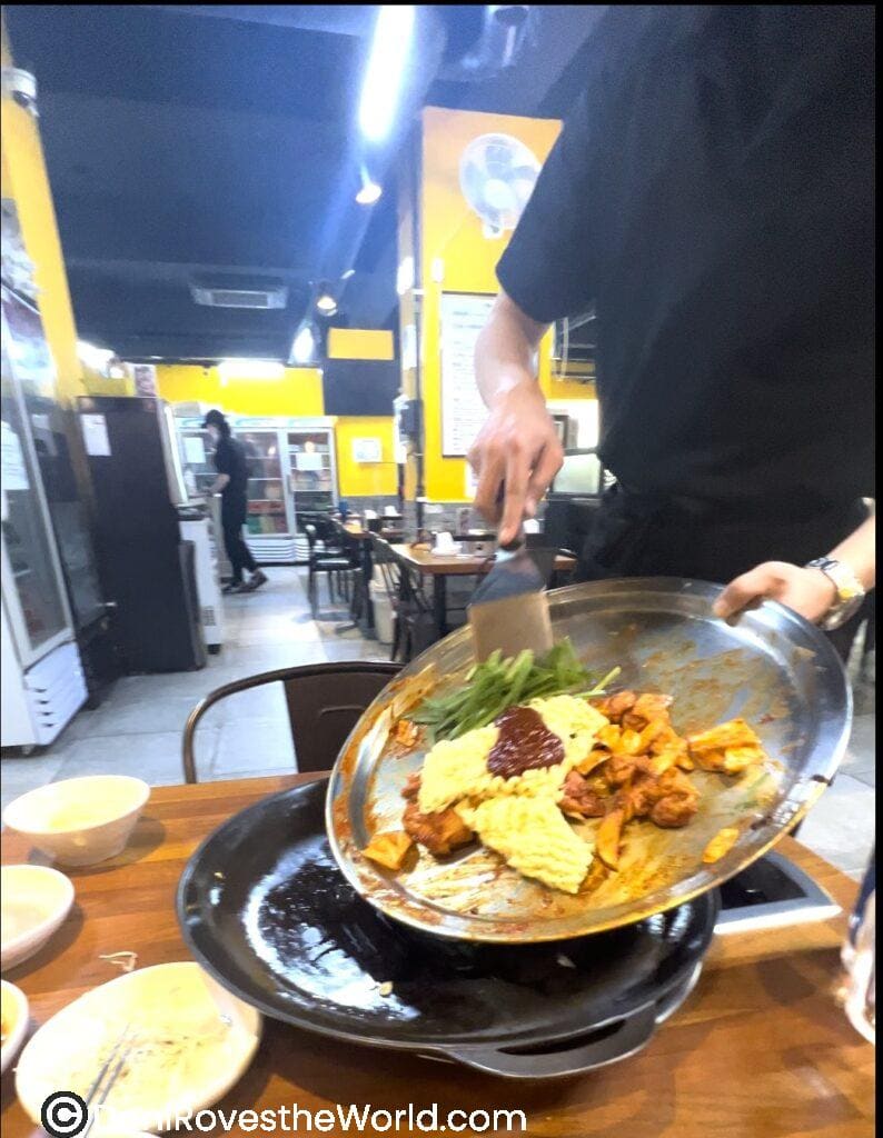 An interactive dining experience in a Hongdae restaurant, with a server grilling marinated chicken (Dakgalbi), vegetables, and noodles at the table, perfect for solo female travelers exploring Korean cuisine. Eating is one of the best things to do in Hongdae.