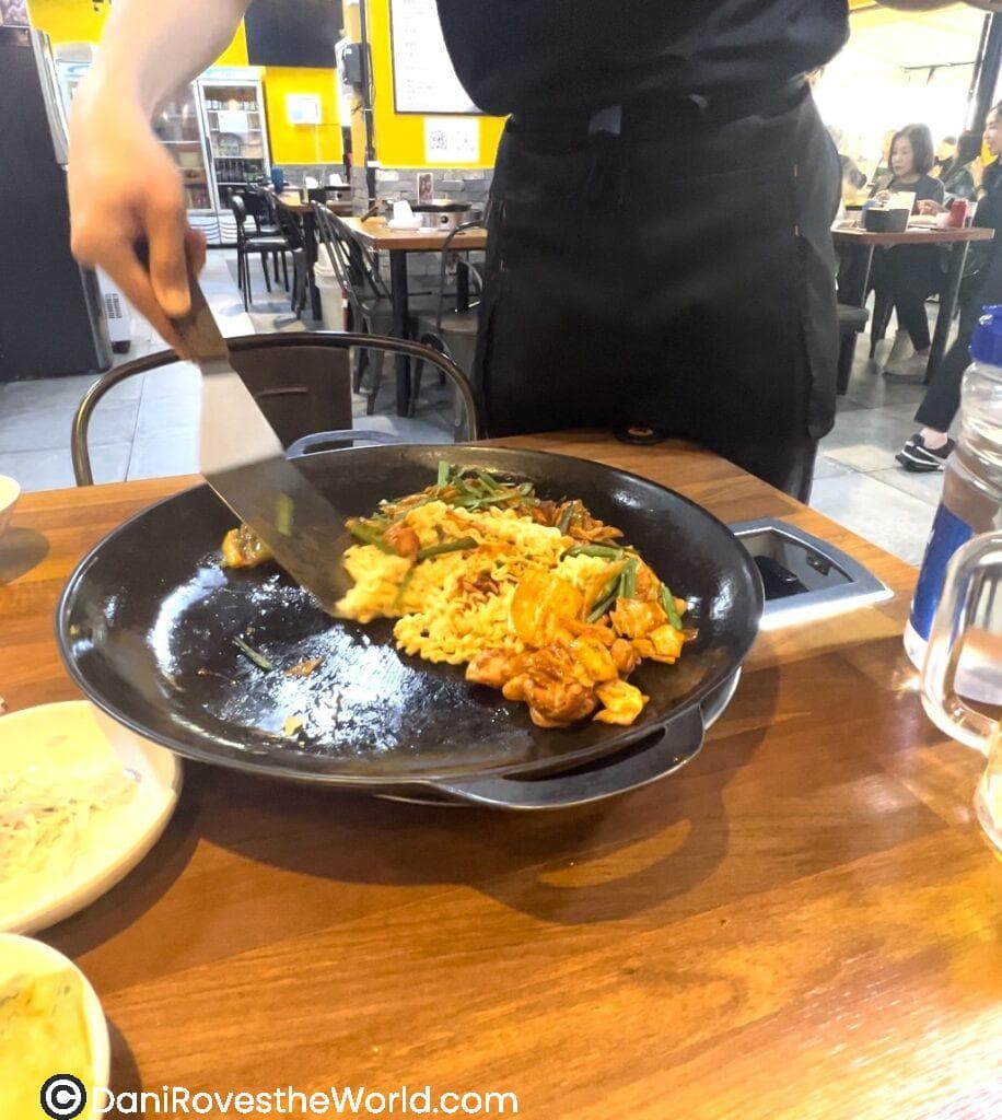 An interactive dining experience in a Hongdae restaurant, with a server grilling marinated chicken (Dakgalbi), vegetables, and noodles at the table, perfect for solo female travelers exploring Korean cuisine.