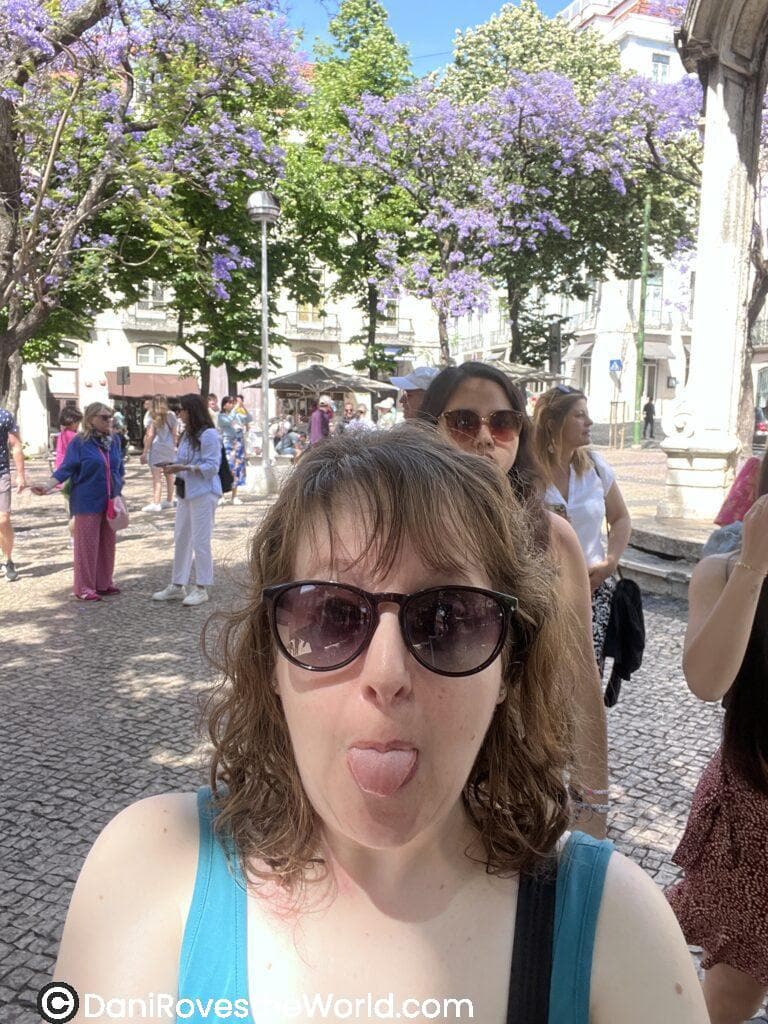 A solo female traveler, Dani, playfully poses in a lively plaza in Lisbon, Portugal with blooming jacaranda trees, highlighting the friendly and relaxed atmosphere that makes this a great destination for solo female travelers.