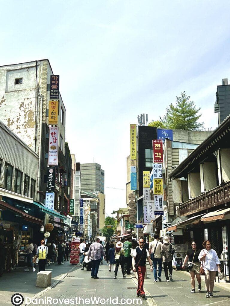 Busy street in Insadong lined with traditional and modern storefronts, featuring signs in Korean hanging from buildings. Pedestrians stroll down the street, enjoying the bustling atmosphere. The mix of old and new architecture reflects Seoul's rich culture. Insadong is a great location to check out on your layover in seoul