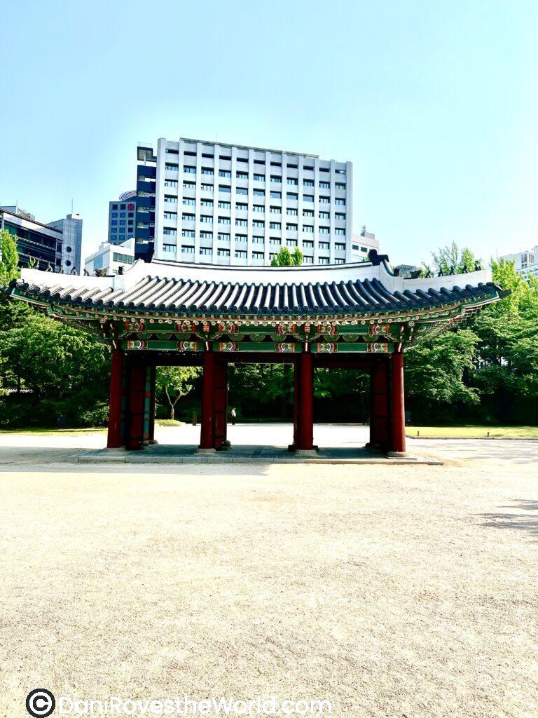 Serene spot to explore during your layover from Incheon airport. Traditional Korean pavilion with ornate detailing, standing in contrast to the modern high-rise buildings in the background. Surrounded by trees and open space, this cultural landmark highlights the blend of history and urban development in Seoul. 