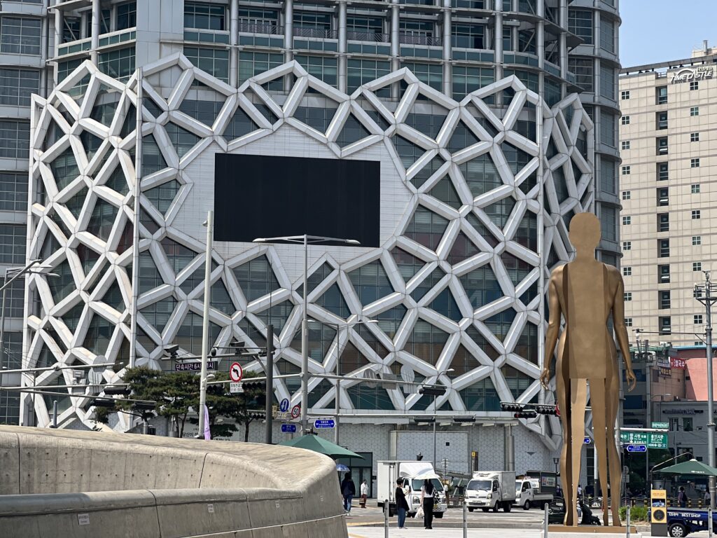 A view of the exterior of Dongdaemun Design Plaza (DDP) in Seoul, with a modern architectural pattern resembling a geometric lattice in the background, alongside a towering bronze human statue, symbolizing the fusion of art and design