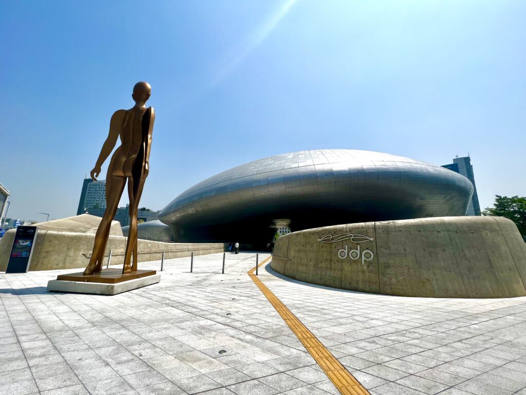 : A bronze statue of a nude human figure stands prominently in front of the futuristic architecture of the Dongdaemun Design Plaza (DDP) in Seoul, a major design and cultural landmark known for its curving, silver facade.