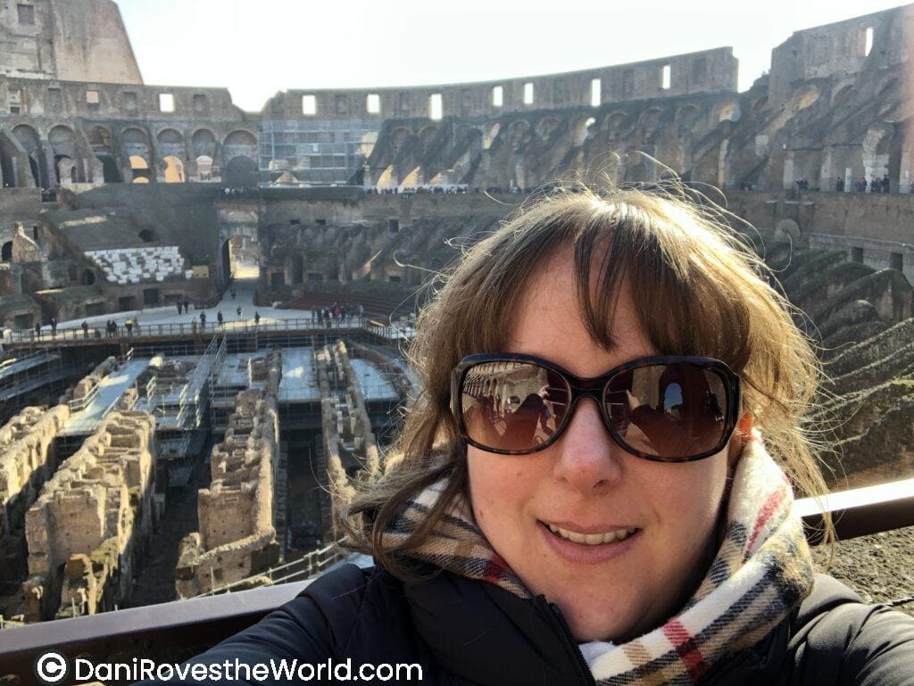 Dani wearing sunglasses and a scarf takes a selfie inside the Colosseum in Rome, capturing the iconic ancient structure in the background. Visiting popular landmarks like the Colosseum during off-peak season is a great budget hack for solo female travelers looking to save money on tickets and avoid large crowds.