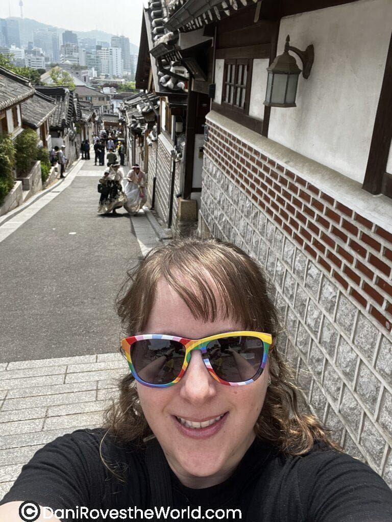 Selfie of a woman wearing rainbow sunglasses in Bukchon Hanok Village, with the traditional Korean rooftops and a distant view of modern Seoul skyscrapers. A blend of history and modernity to experience within 24 hours in Seoul.