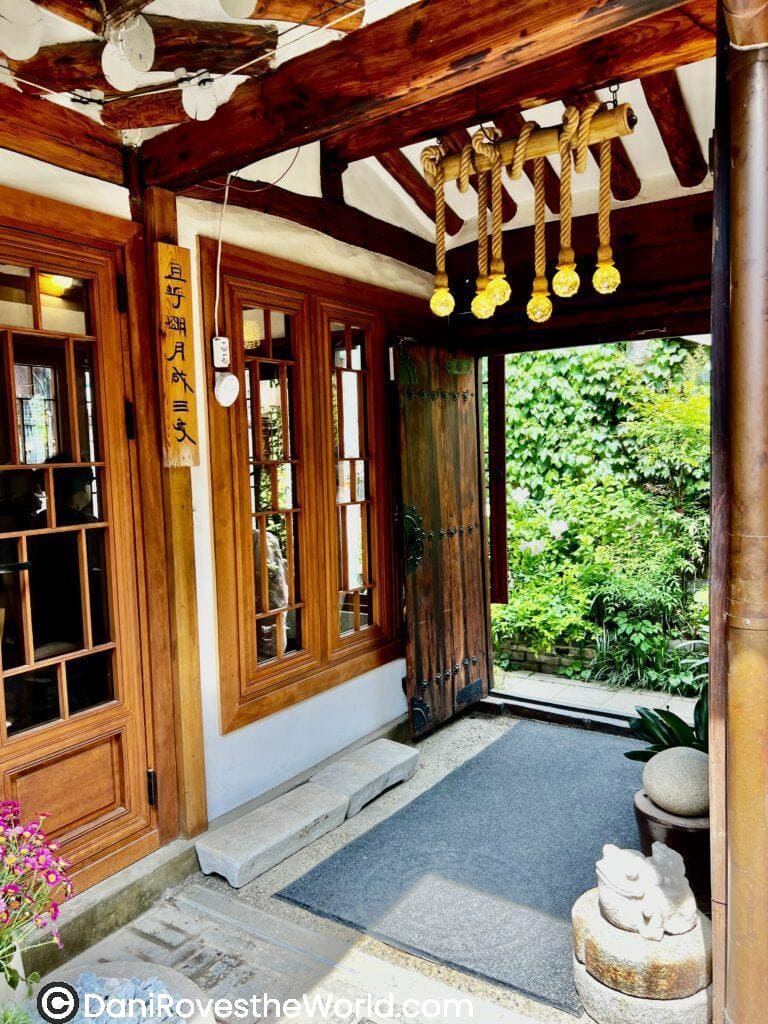 The entrance of a traditional Korean hanok, featuring wooden doors and a lush garden, creating a calming and inviting space for relaxation