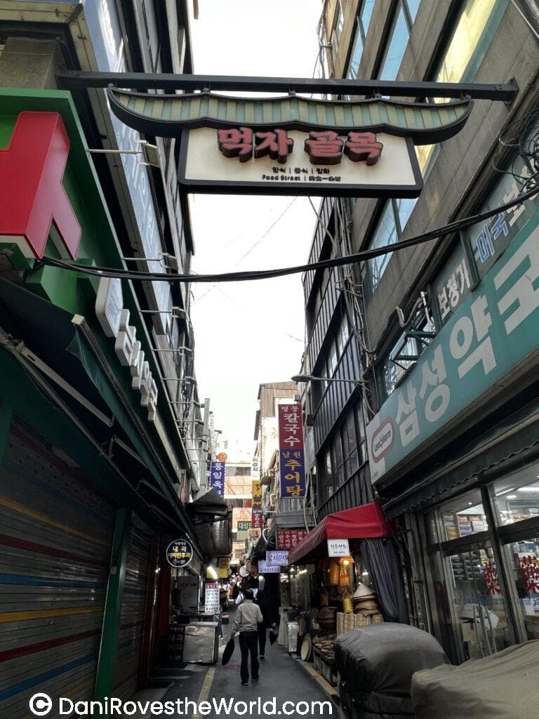 A narrow alleyway in Korea, with a large sign overhead reading "Food Street" in both English and Korean. The alley is filled with small shops, some with colorful awnings, and a few people walking down the path. The area gives a cozy and bustling vibe typical of a local market district.