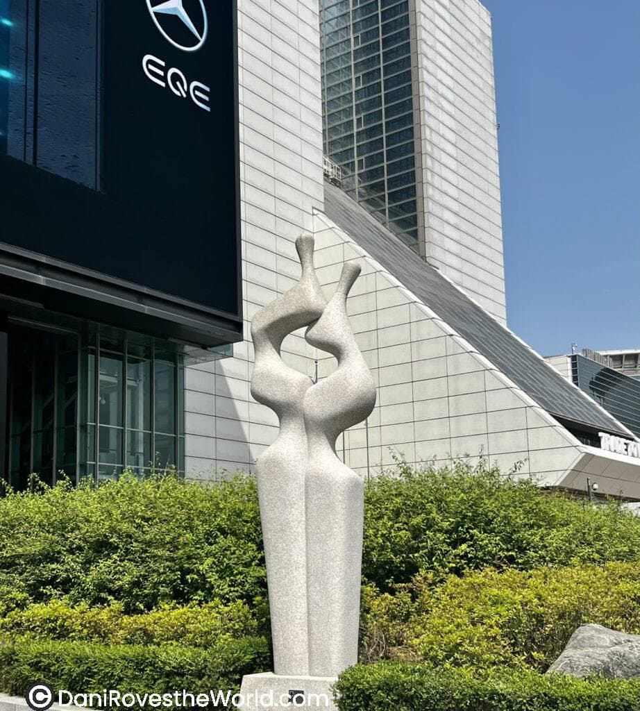 A modern sculpture in front of a high-rise building in an urban setting. The abstract sculpture features two intertwined shapes, made of light-colored stone. In the background, a black Mercedes-Benz sign with the "EQE" logo is visible on the building.