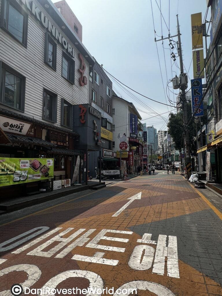 A street scene in Korea with shops and restaurants lining both sides. The pavement features an arrow pointing forward, and Korean text painted on the street. People can be seen walking down the road, with visible signs for local businesses on either side. The sky is bright and sunny.