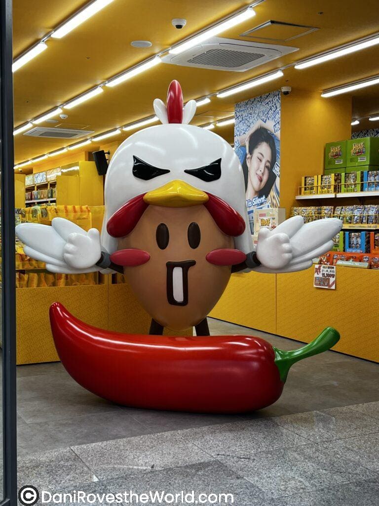 A fun and colorful statue of a cartoon chicken character holding a large red chili pepper, located inside a Myeongdong snack shop. This playful spot is a popular stop for visitors seeking quirky photo opportunities and local treats as part of their things to do in Myeongdong itinerary.