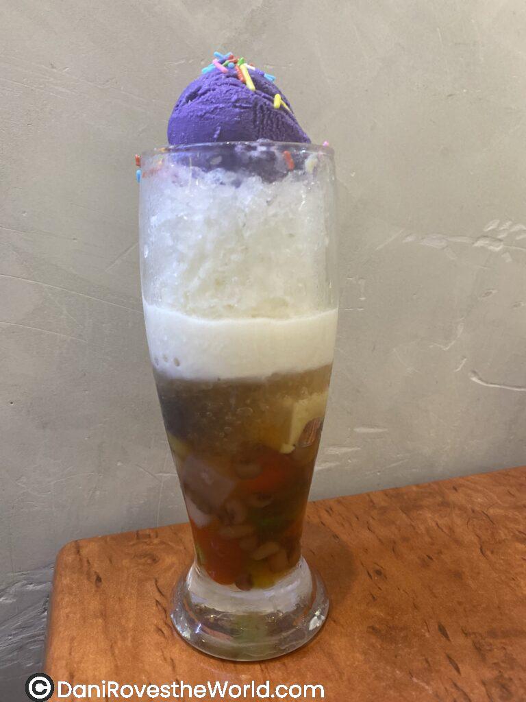A tall glass of halo-halo, a popular Filipino dessert, topped with vibrant purple ube ice cream and colorful sprinkles. The layers of crushed ice, milk, and mixed ingredients at the bottom create a refreshing and unique experience for solo female travelers exploring Filipino cuisine abroad.