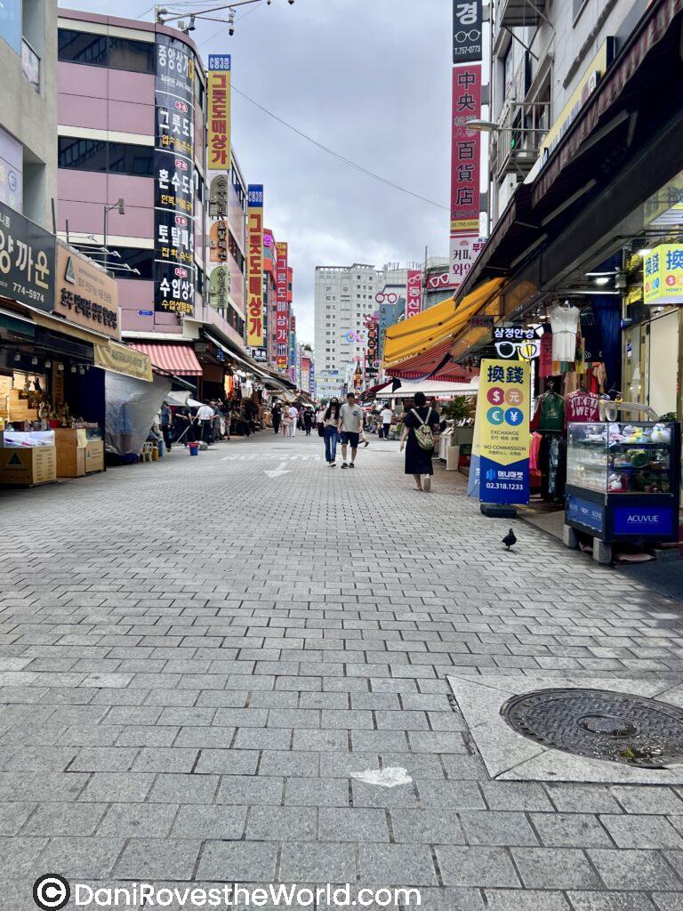 A urban street scene in Korea, lined with shops displaying Korean signs and banners. People stroll down the wide paved road, flanked by market stalls and colorful awnings. The sky is overcast, and the street is framed by tall, modern buildings in the background.
