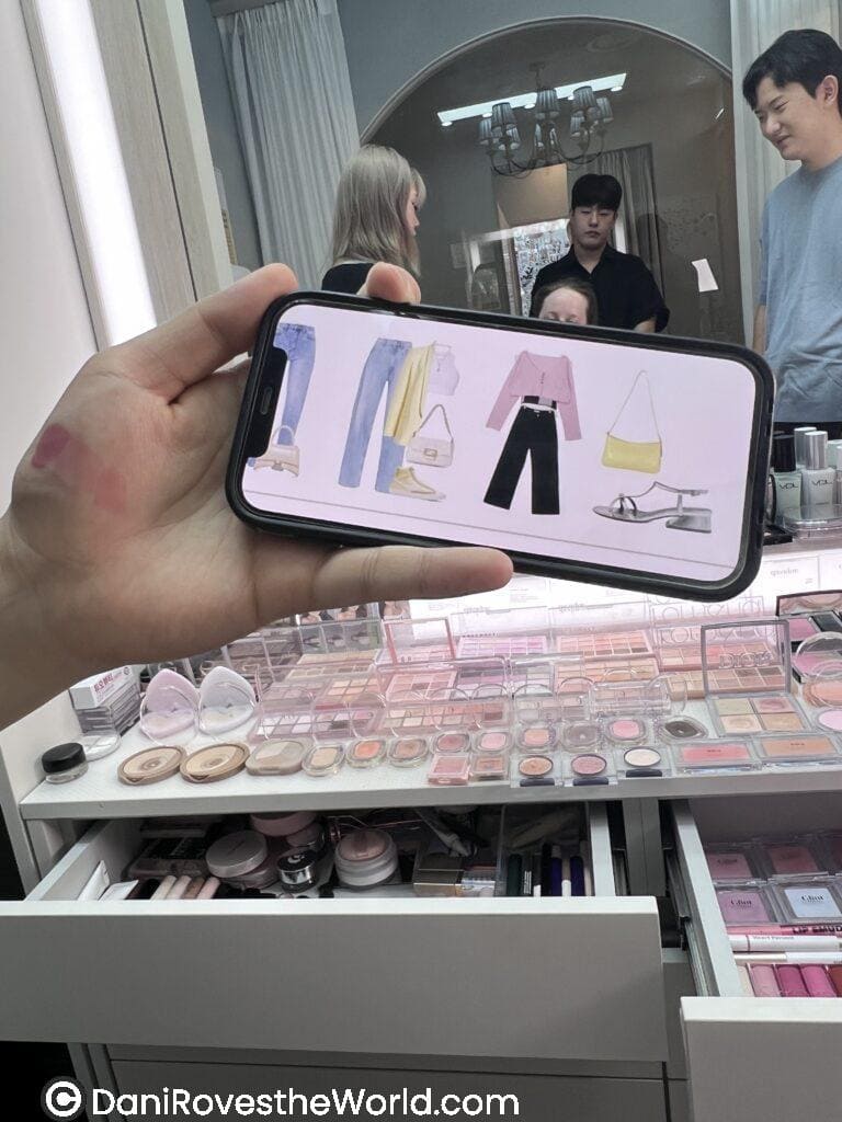 A hand holding a phone showing fashion outfits, in front of a makeup counter in a Seoul beauty shop. One of the steps in getting your color analysis done in Hongdae, Seoul. 