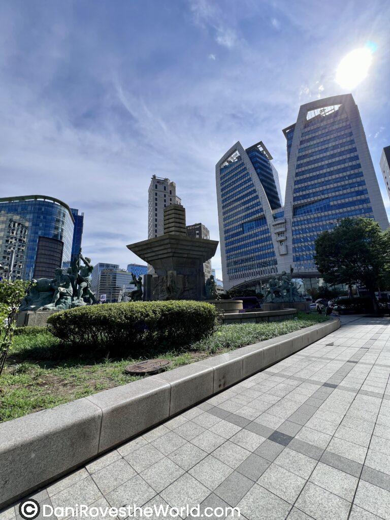 A view of modern skyscrapers and a public fountain in Seoul, South Korea, under a bright blue sky. The scene captures the blend of nature and urban architecture typical in the city