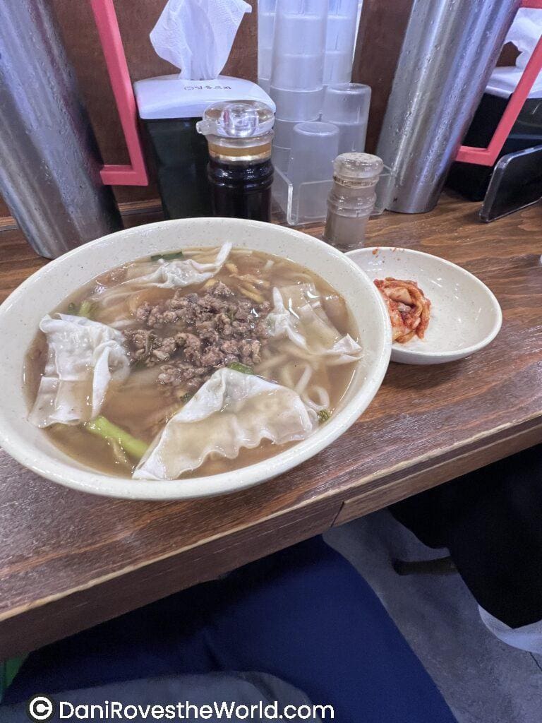 A warm, comforting bowl of Korean dumpling noodle soup served with a side of spicy kimchi, providing solo female travelers with a hearty meal option during their exploration of Asian street food and local eateries.