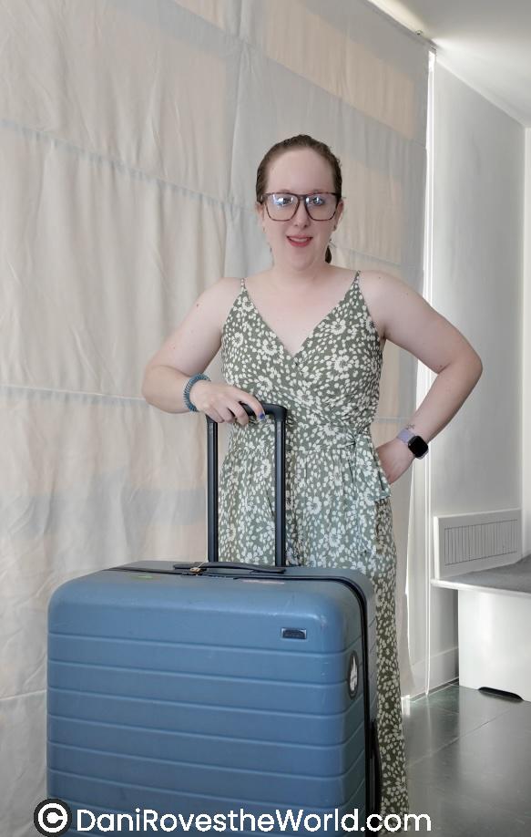  A person wearing glasses and a green floral dress, standing with a blue hard-shell suitcase. The person is smiling and standing indoors, ready for travel. 