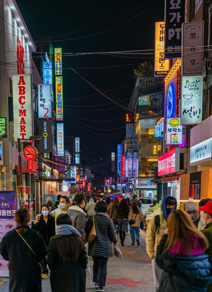 A busy street in Seoul at night, filled with people walking amidst bright neon signs and storefronts. The scene showcases the city's lively nightlife and pedestrian culture