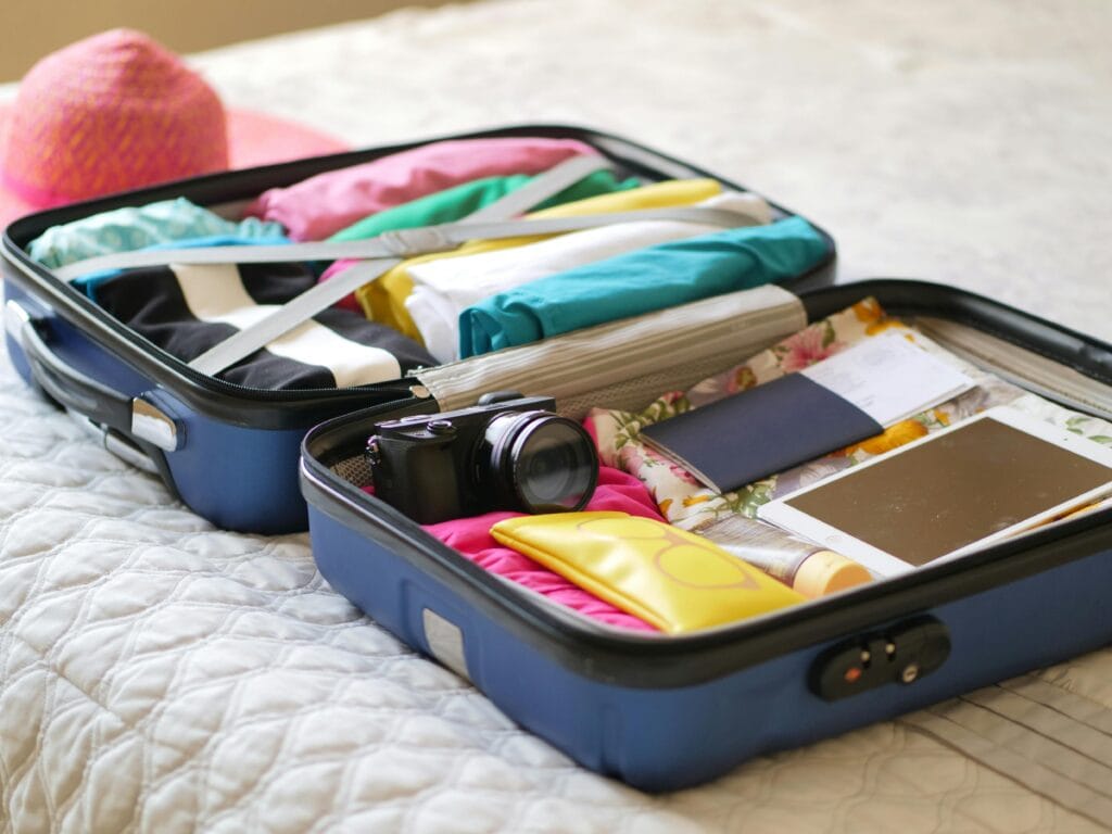 An open suitcase on a bed, neatly packed with colorful clothes, a camera, a tablet, a passport, and travel documents. A pink sunhat is partially visible beside the suitcase, suggesting preparation for a trip.