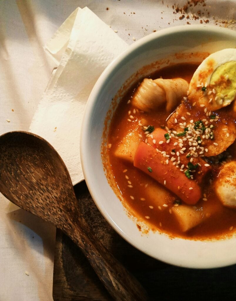 A bowl of spicy tteokbokki, a popular Korean street food, topped with sesame seeds, fish cakes, and a boiled egg. Sampling traditional street foods like tteokbokki is one of the must-try things to do in Myeongdong for food lovers
