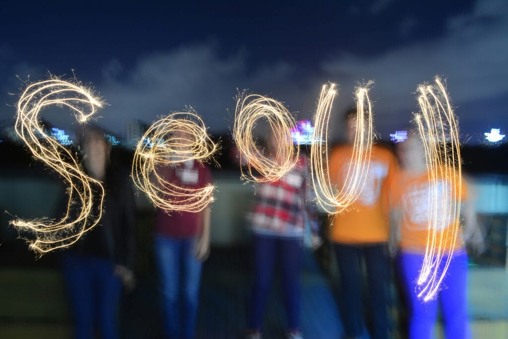 Five people stand blurred in the background while writing the word 'Seoul' with sparklers in the air, symbolizing the vibrant and creative spirit of the city at night