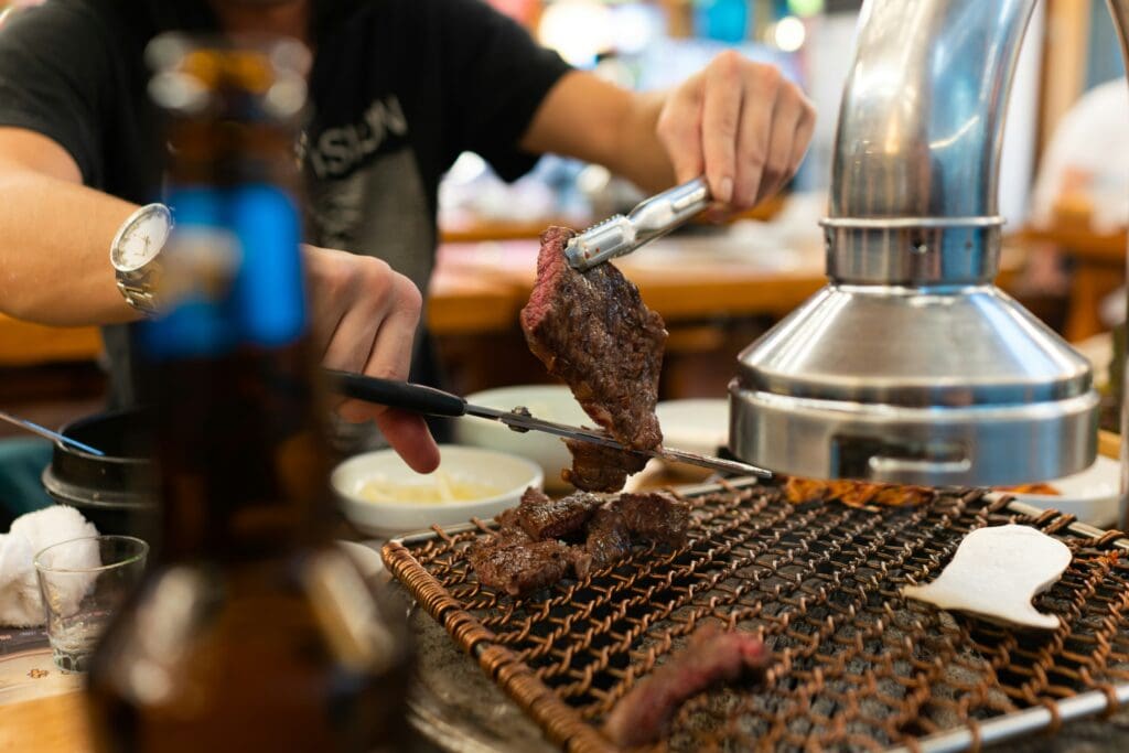 A person grilling marinated meat over an open charcoal grill at a Korean BBQ restaurant in Seoul. The scene captures the hands-on dining experience with juicy beef on the grill.