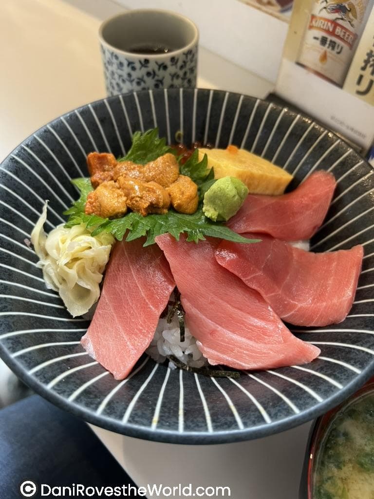 A bowl of fresh sushi rice topped with slices of raw tuna, sea urchin (uni), tamago (Japanese omelet), wasabi, pickled ginger, and shiso leaves, served in a black, striped bowl. A cup of tea and a Kirin beer advertisement are visible in the background.