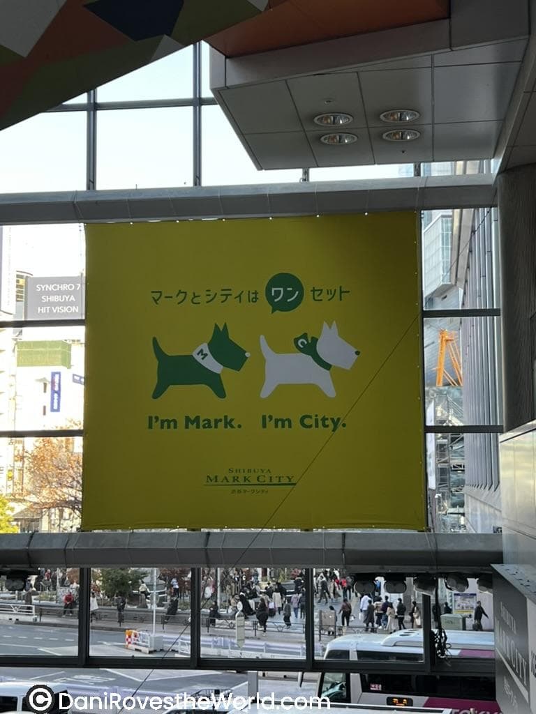 A large yellow banner inside Shibuya Mark City featuring two cartoon dogs: one green and one white, with text in English that reads, “I’m Mark. I’m City.” Japanese text above mentions they are a “set,” and a crowd of people and city streets are visible through the windows below.
