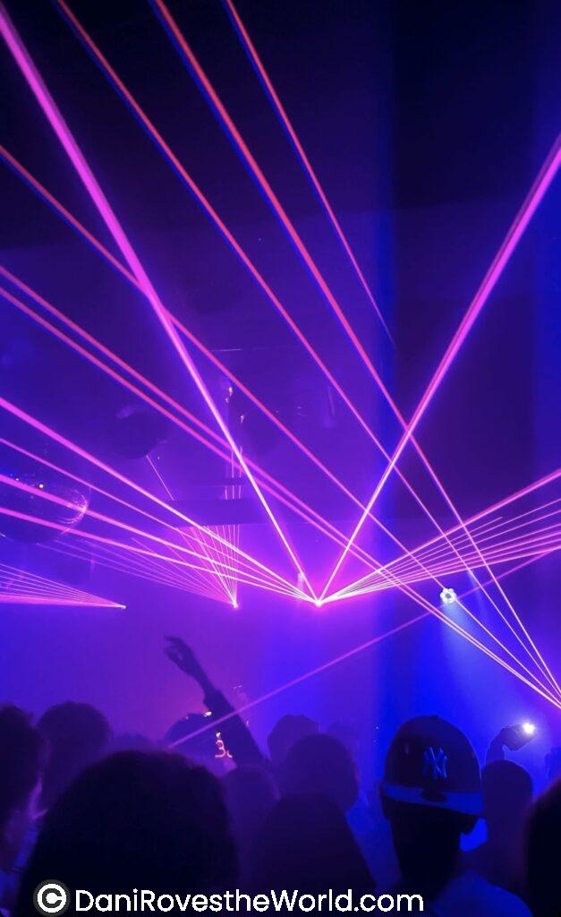 A vibrant nightclub scene with a crowd of people silhouetted against bright purple and pink laser lights cutting across the dark venue. One person’s hand is raised in the air, and another wears a dark cap with a visible New York Yankees logo.