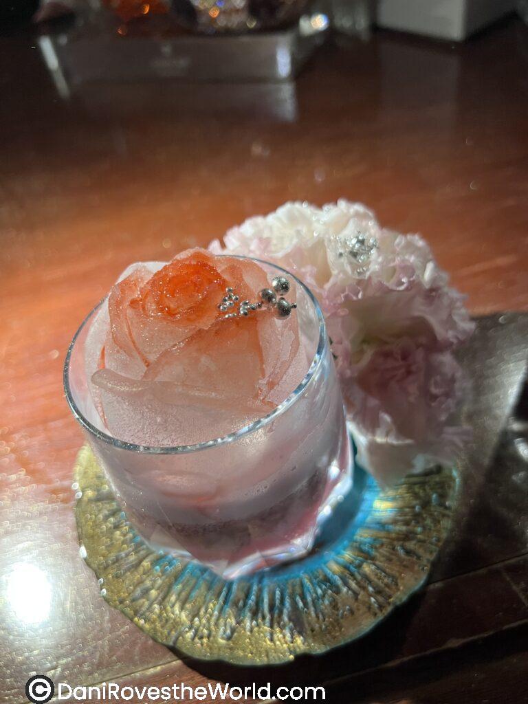 Beautiful rose cocktail with glitter. The ice cube is shaped like a rose and there is a flower on the tray that is serving the drink.