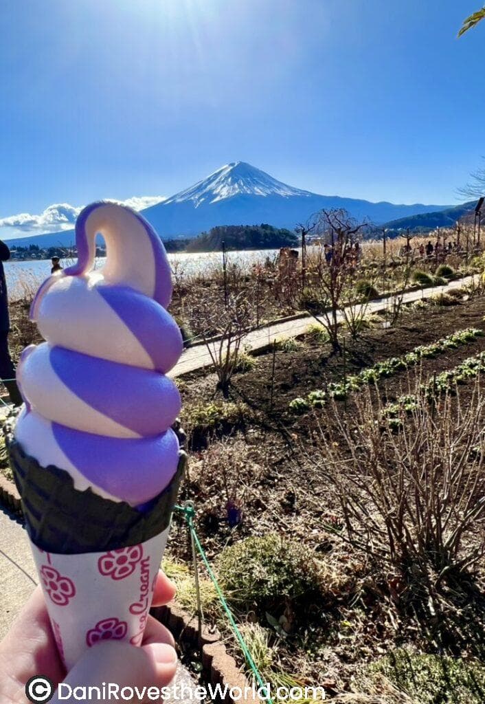 A vibrant lavender and white swirl ice cream cone is held in the foreground, with Mount Fuji majestically rising in the background under a sunny sky, surrounded by a garden pathway.
