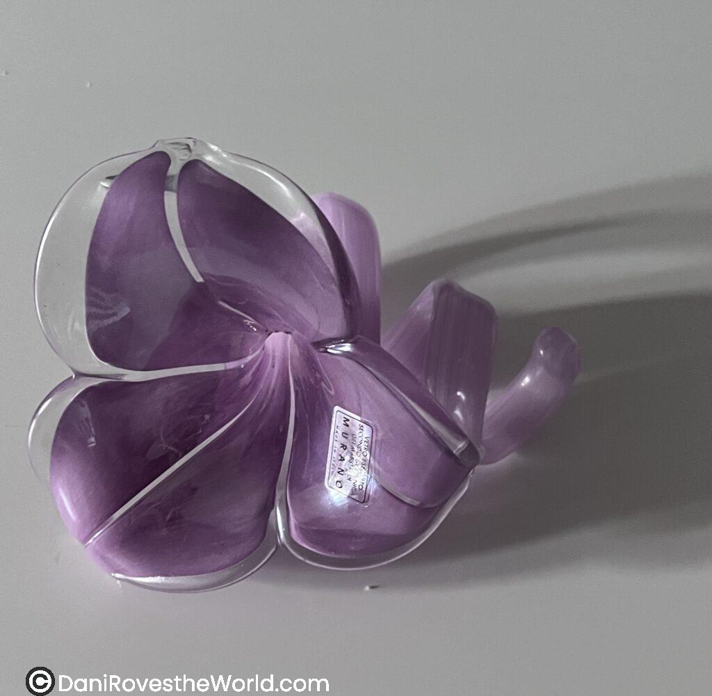 A glass sculpture of a purple flower, labeled "Murano," with a glossy, translucent finish and subtle swirling patterns