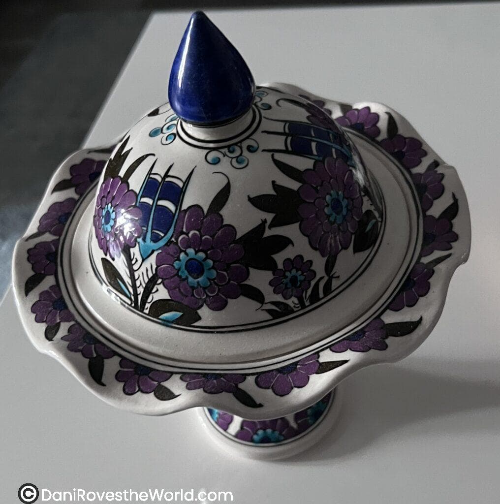 A decorative lidded ceramic bowl with intricate purple and blue floral patterns, accented by a deep blue handle on top of the lid.