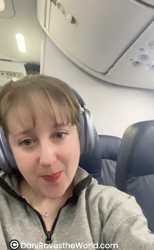 A woman wearing headphones, seated in an airplane, takes a selfie. She has a slight smile, with a soft lighting highlighting the cabin interior behind her.